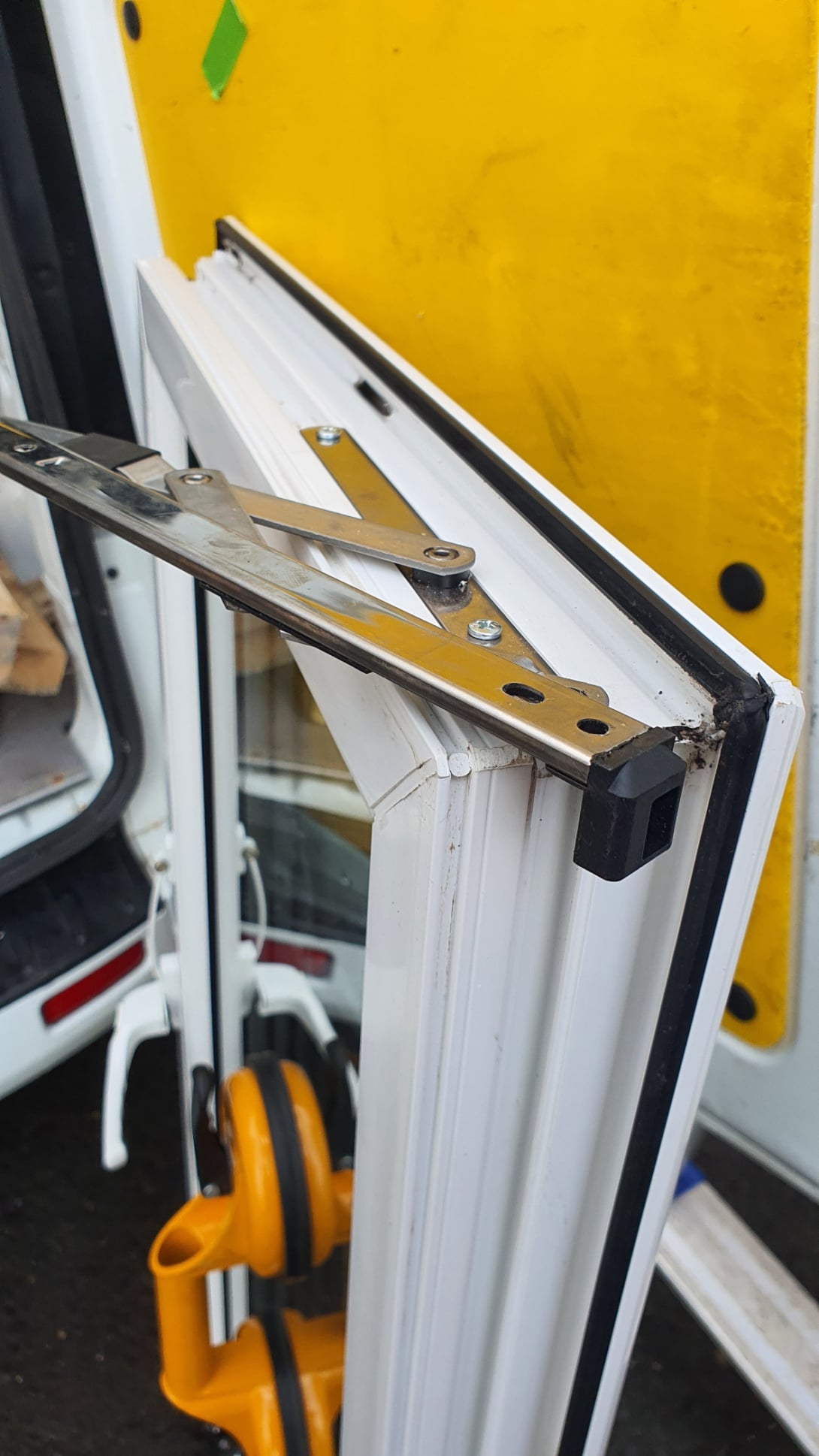 “Close‑up of a uPVC window hinge mechanism during repair, showing the metal friction stay, screws and corner buffer with tools visible in the background.