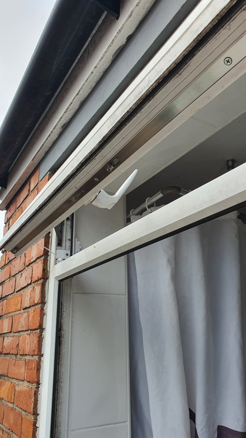 A white uPVC tilt‑and‑turn window is shown partially open inward, revealing the hinge and tilt mechanism on the side of the sash. The window is set in a brick wall, with indoor piping, tiles, and a curtain visible in the background.