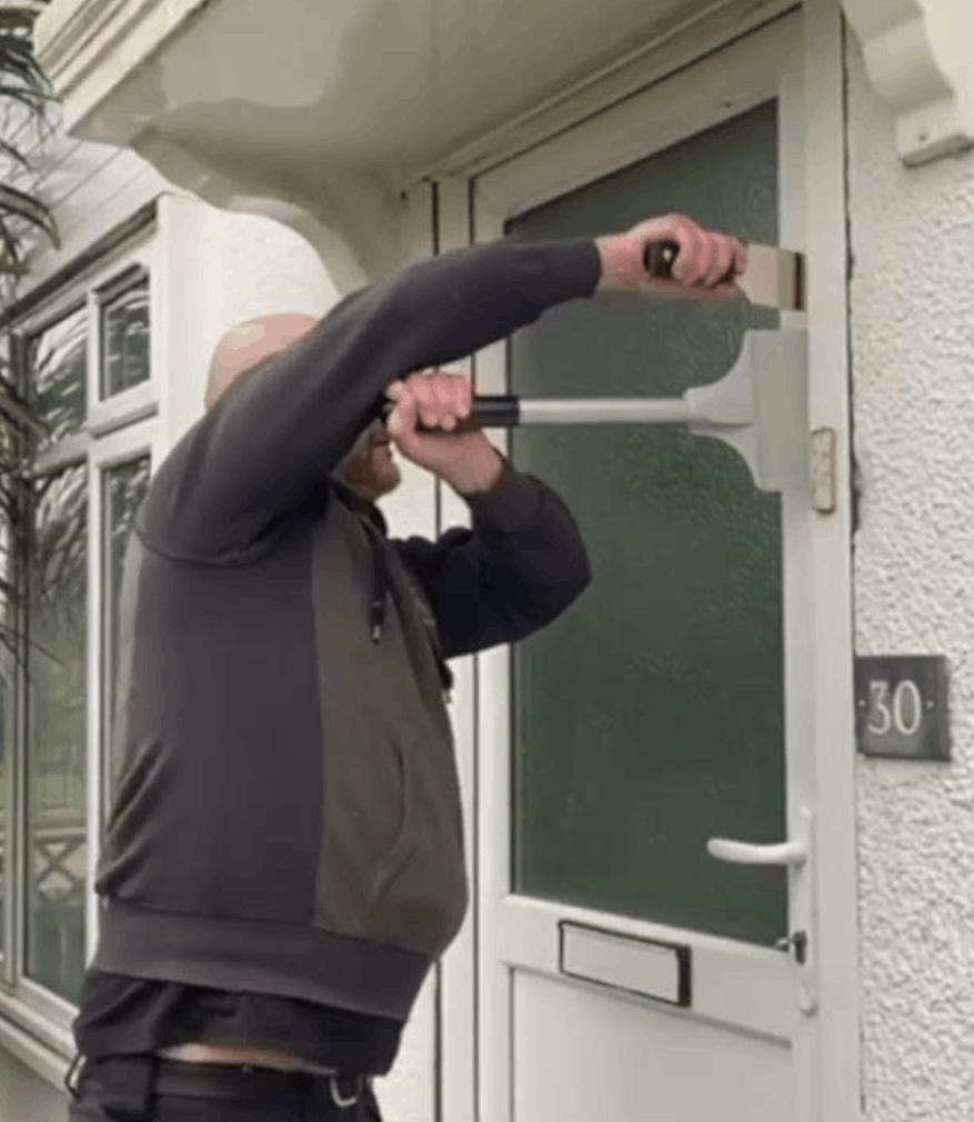 The image shows someone using a professional tool, probably a door spreader, to safely open a door. The door has the number "30" and a frosted glass panel. The person appears to be prying near the lock area.