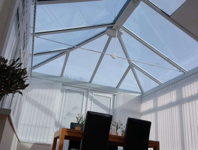 Interior view of a conservatory roof with triangular glass panels and a central hub fitted with clamps during repair work, with a ladder visible outside the window.
